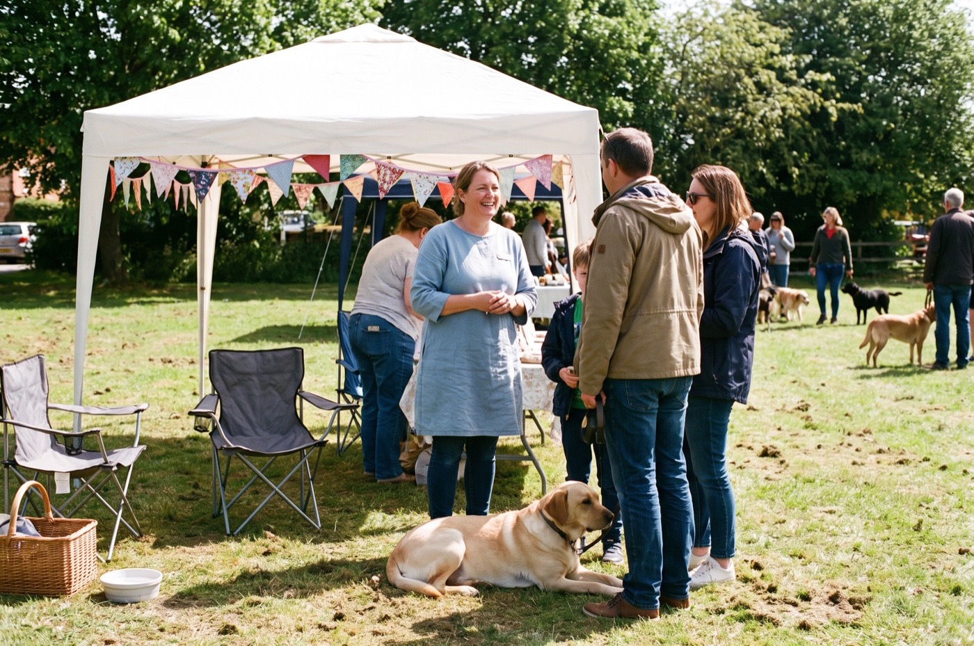 A groomer chats with a family beside a white gazebo with bunting at a sunny village dog show, their Labrador lying in the grass
